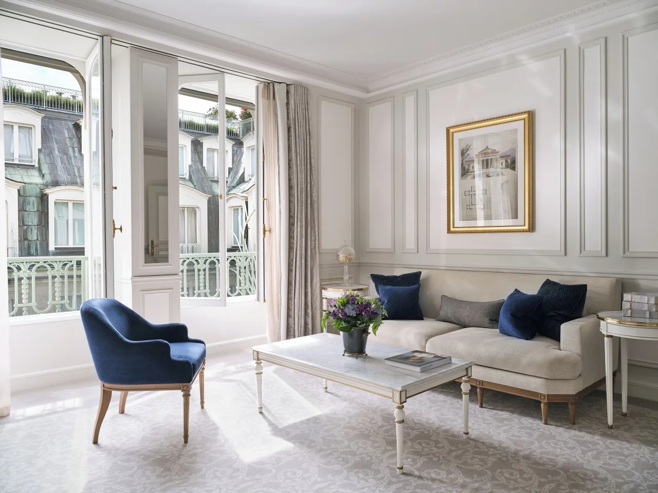 Un salon clair à boiseries, un fauteuil de velours bleu marine, un canapé blanc aux coussins bleus et des portes-fenêtres sur un garde-corps