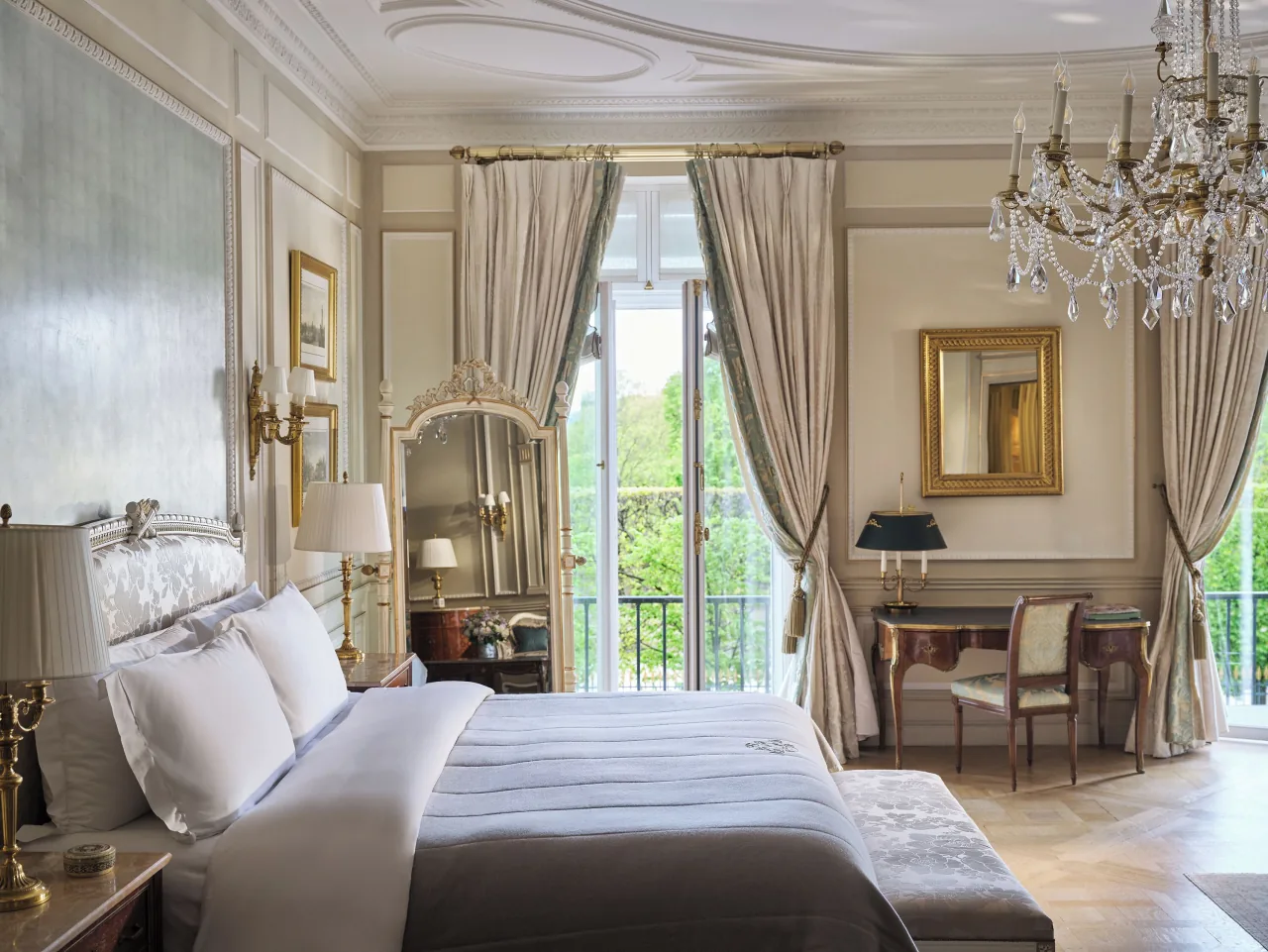 Une chambre, un lit blanc, un lustre de cristal, un miroir doré, un bureau et des portes-fenêtres ouvertes sur des arbres