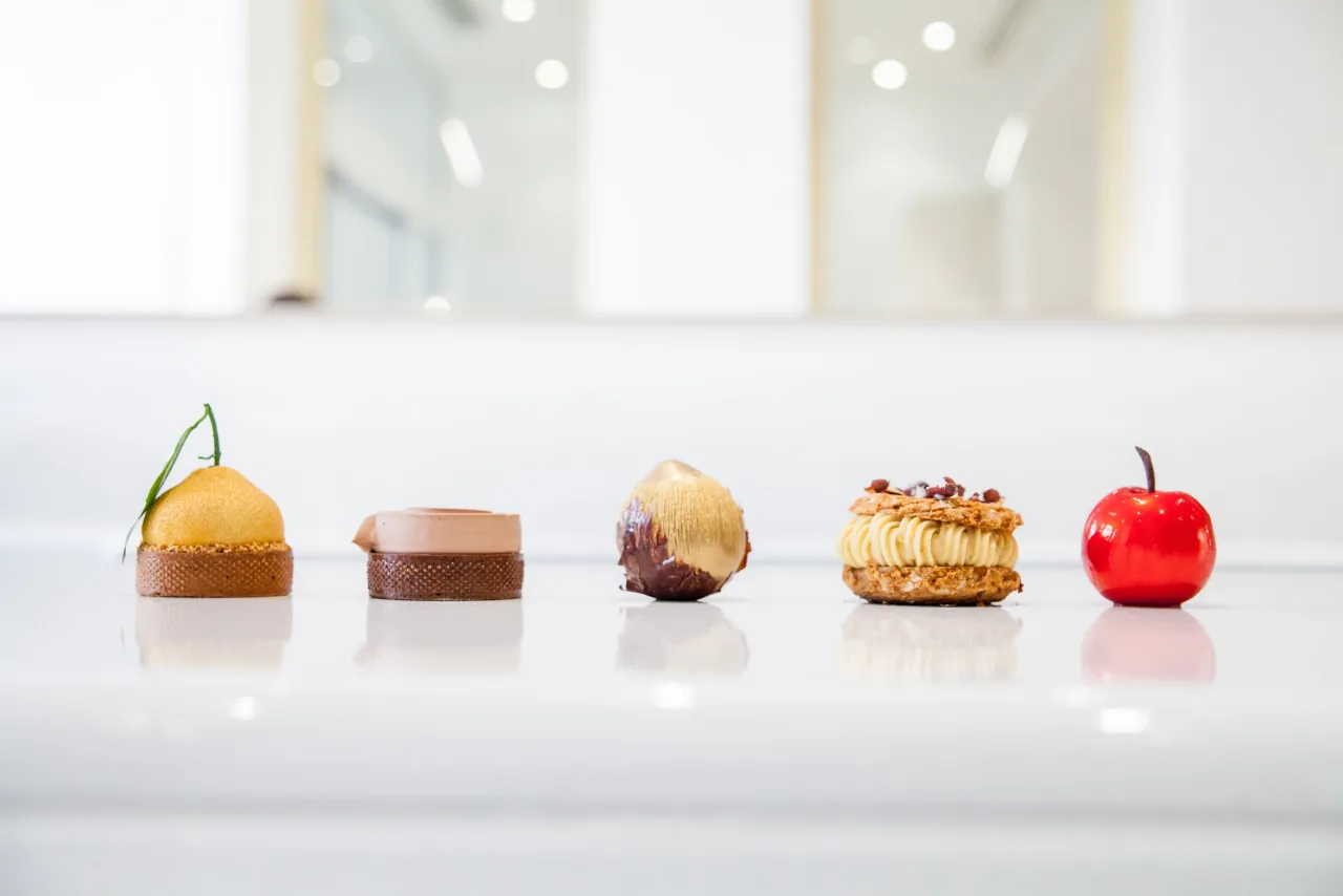 Cinq pâtisseries alignées sur un comptoir blanc, dont une en forme de pomme rouge