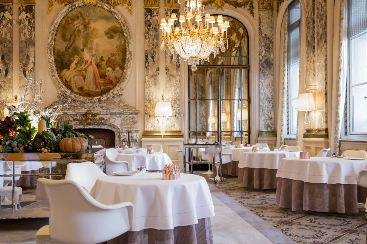 Une salle à manger aux boiseries dorées, un médaillon peint au-dessus d’une cheminée de marbre, des lustres de cristal et des tables rondes aux fauteuils blancs