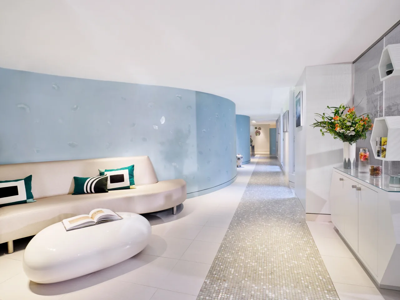 Un salon blanc, un canapé incurvé, une table en forme de galet, un mur courbe bleu pâle et un couloir de mosaïque