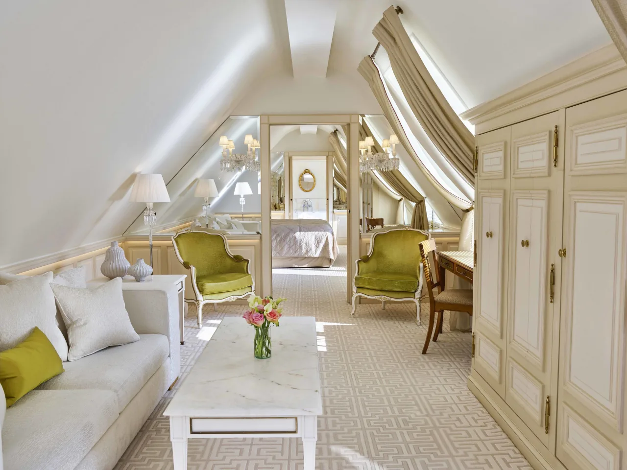 Un salon crème sous des combles mansardés, des fauteuils verts, un canapé blanc, une table de marbre et une chambre au fond