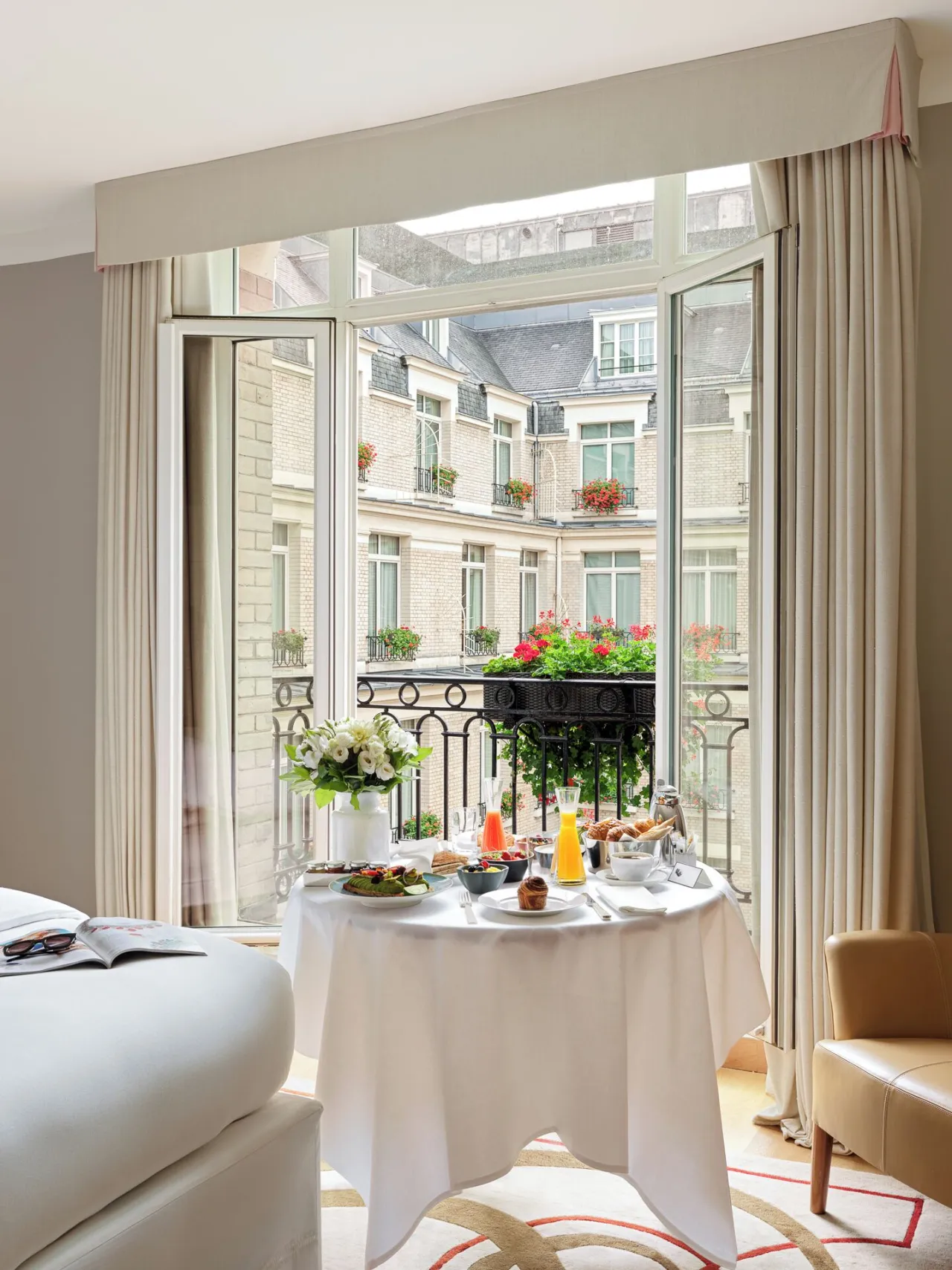 Un petit-déjeuner servi près d’une porte-fenêtre ouverte sur la cour, jardinières fleuries aux balcons d’en face