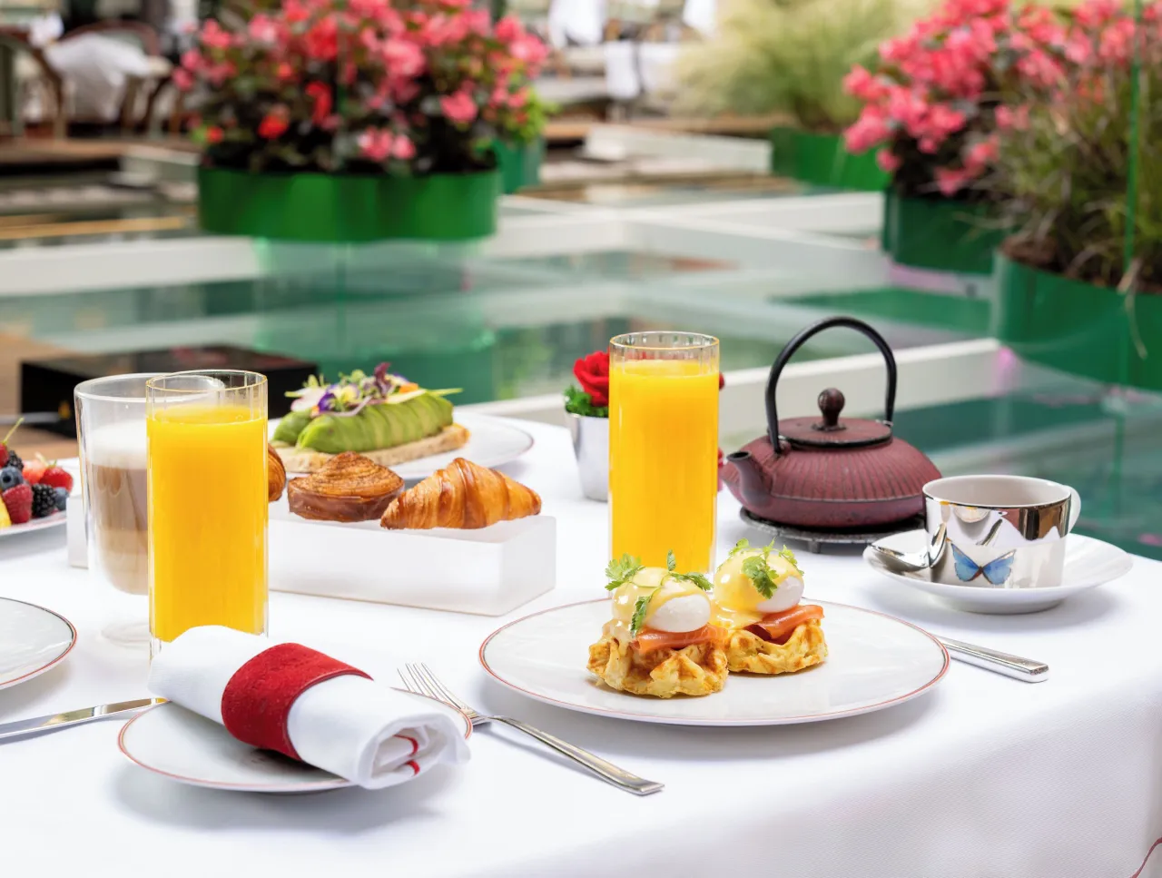 Une table de petit-déjeuner, jus d’orange, viennoiseries, œufs et théière en fonte, fleurs à l’arrière-plan