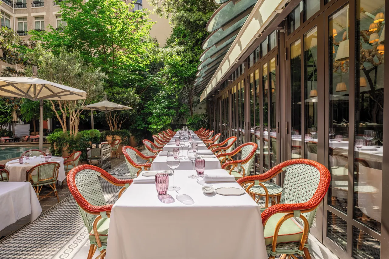 Une longue table dressée sur une terrasse sous les arbres, chaises en rotin rouge et portes vitrées sur un côté