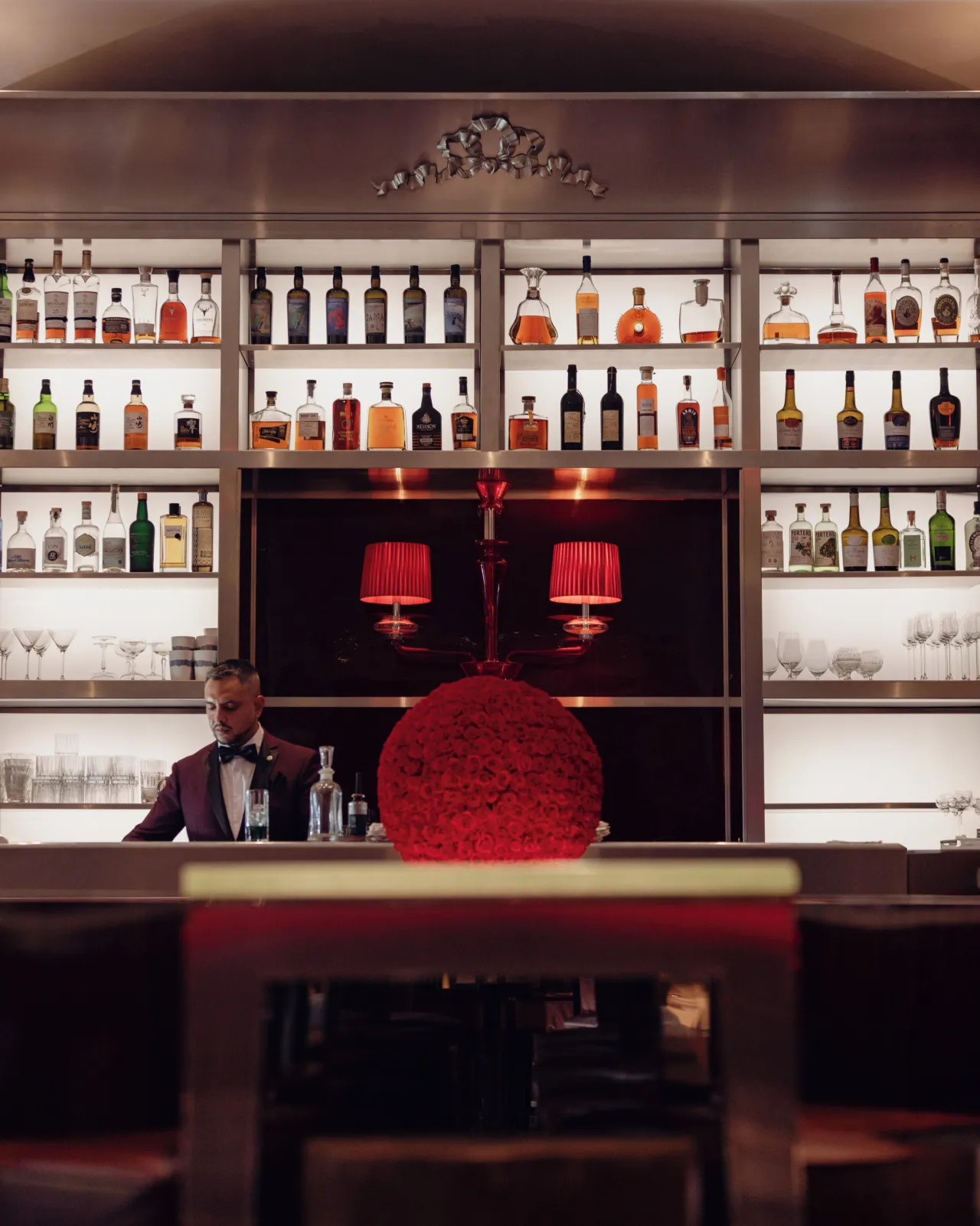 Un bar, étagères de bouteilles, lampes rouges et barman derrière le comptoir