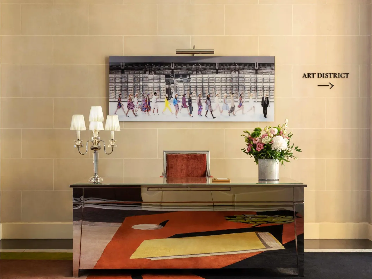 Un tableau de personnages sur un mur clair, au-dessus d’une console en miroir, avec des fleurs et un panneau « Art District »