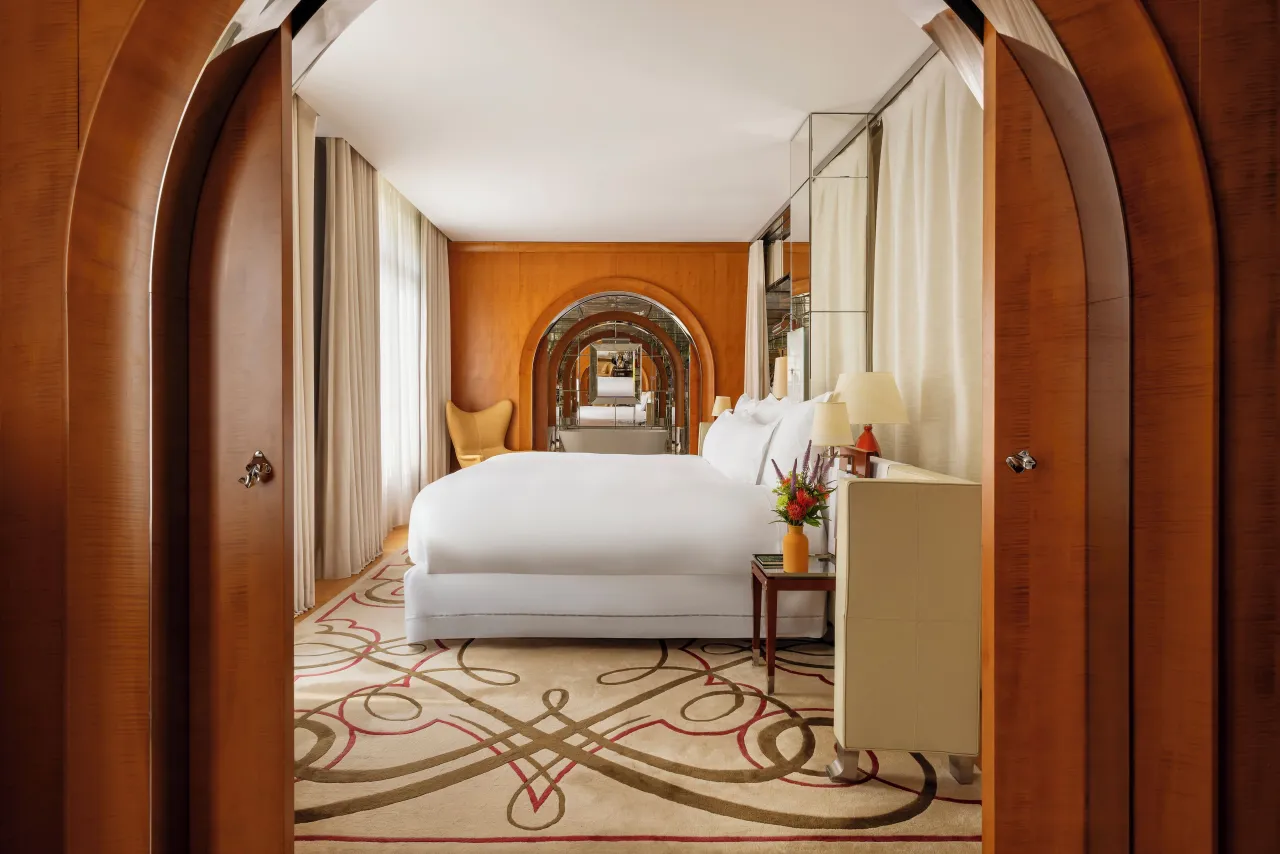 Une chambre vue par deux portes de bois cintrées ouvertes, lit blanc et tapis à motifs