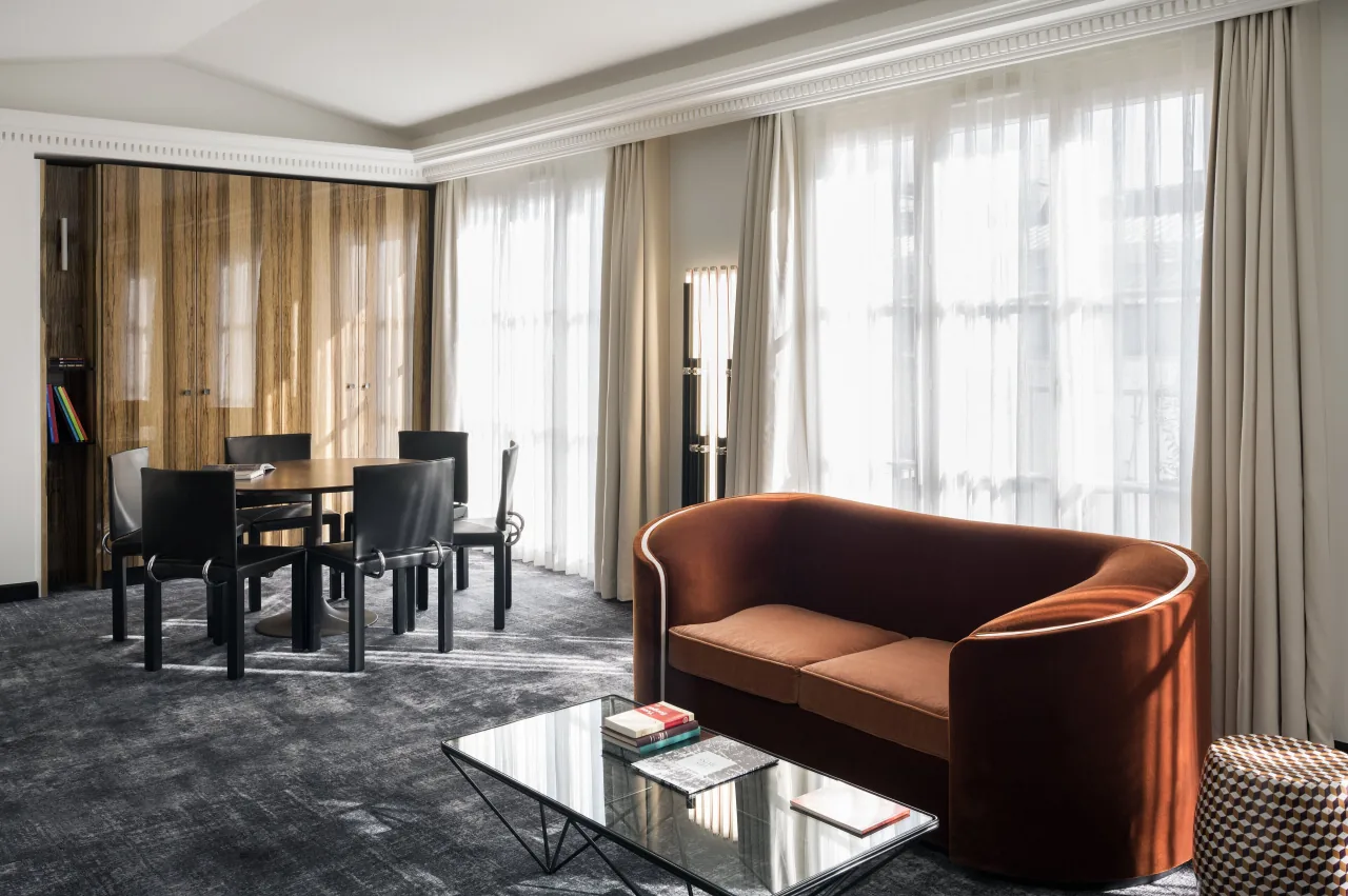 A lounge with a rust velvet sofa, a glass coffee table and a dining table, wood panelling and tall curtains