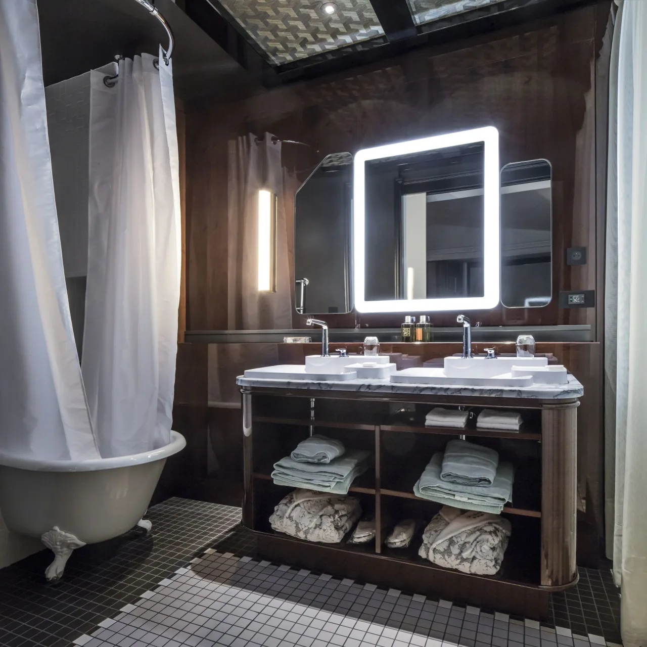 A bathroom with a claw-foot bathtub, a double washbasin over shelves of towels and a lit mirror