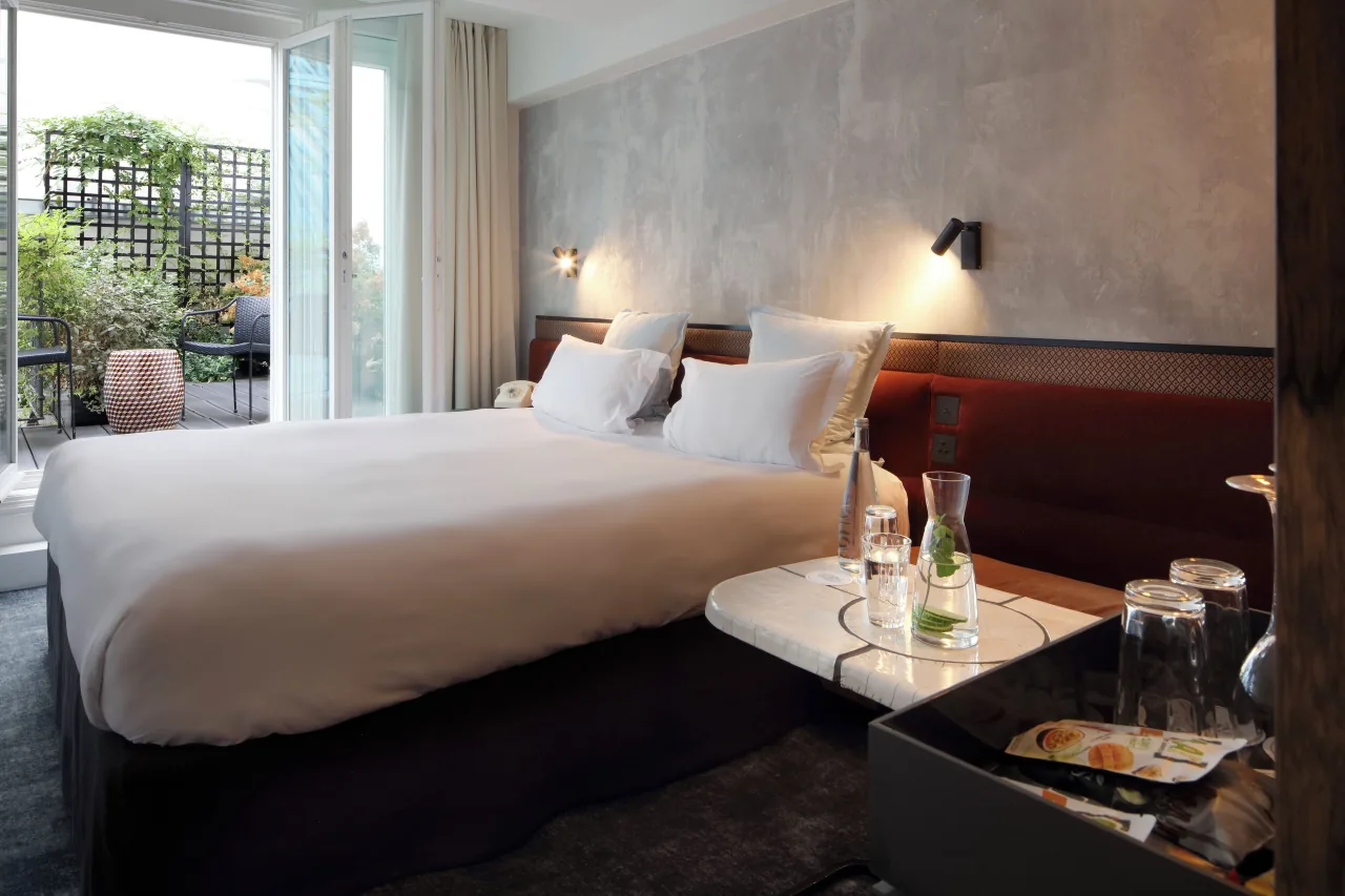 A bedroom with a large white bed and a grey wall, its window open onto a planted terrace