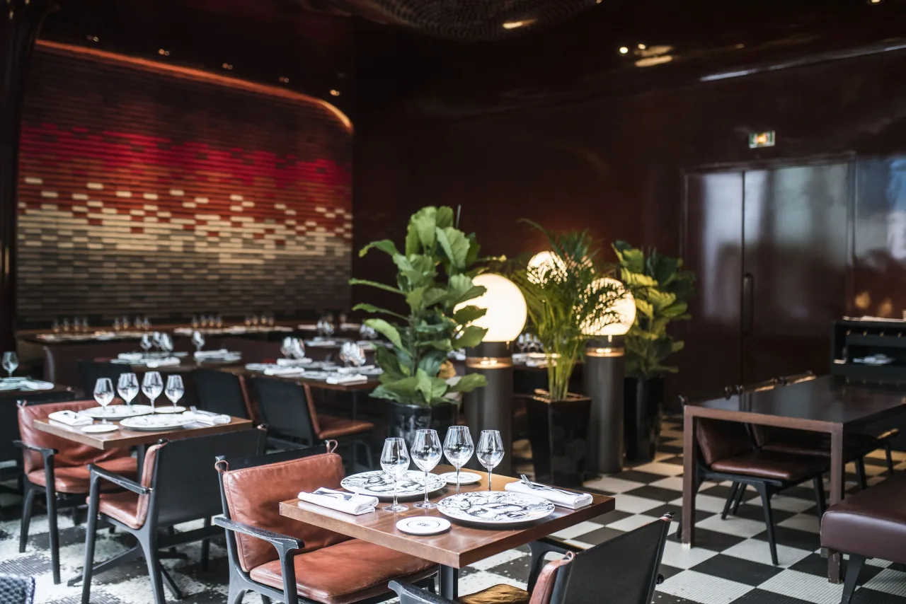 A restaurant with set tables, leather chairs, plants and a checkerboard floor under dark lacquered walls