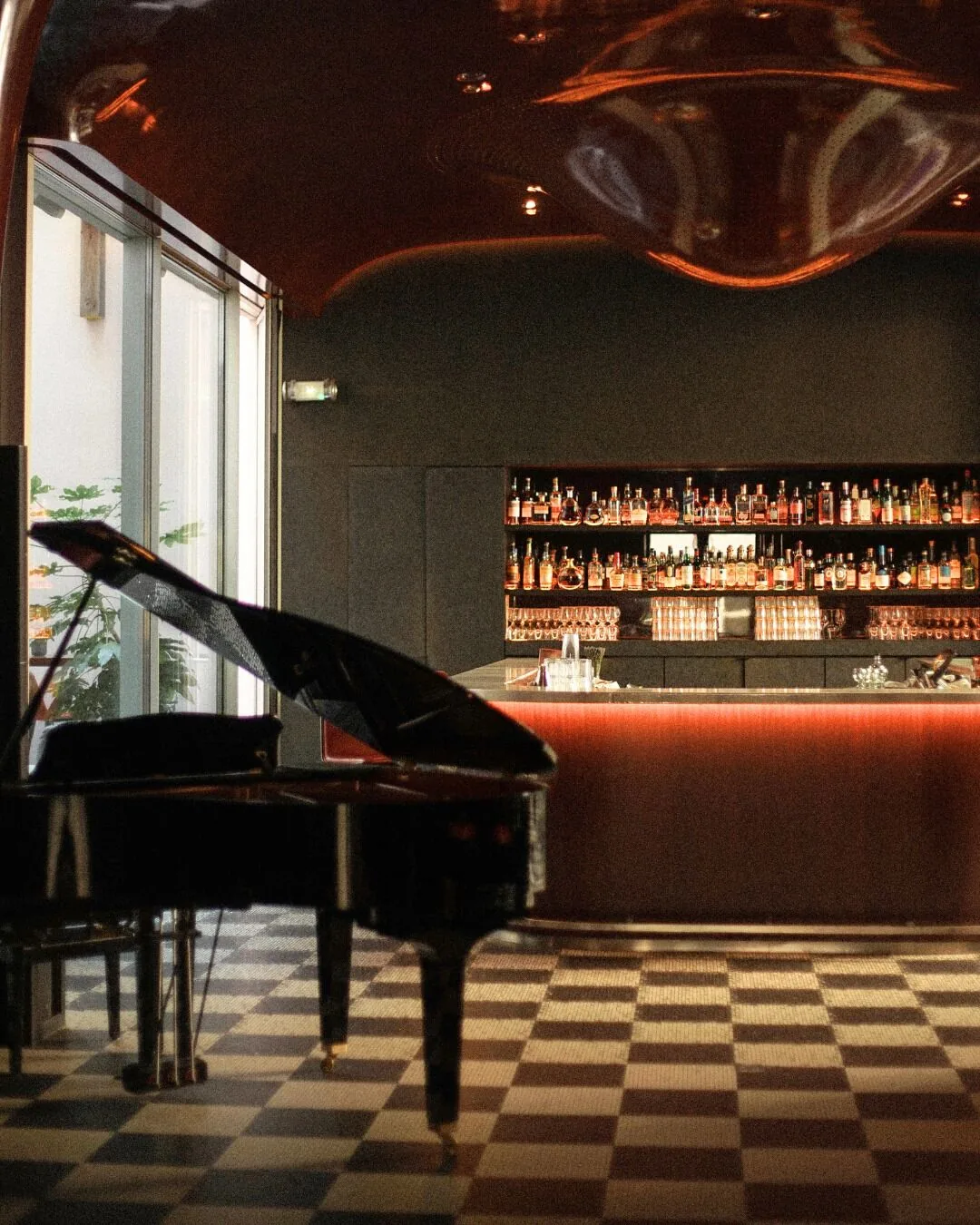 A grand piano in front of a bar with lit shelves, on a checkerboard floor