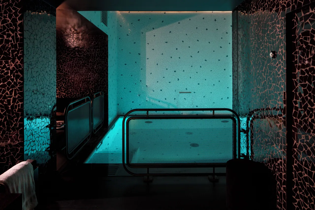 An indoor pool lit in turquoise, with dark mosaic walls and chrome handrails