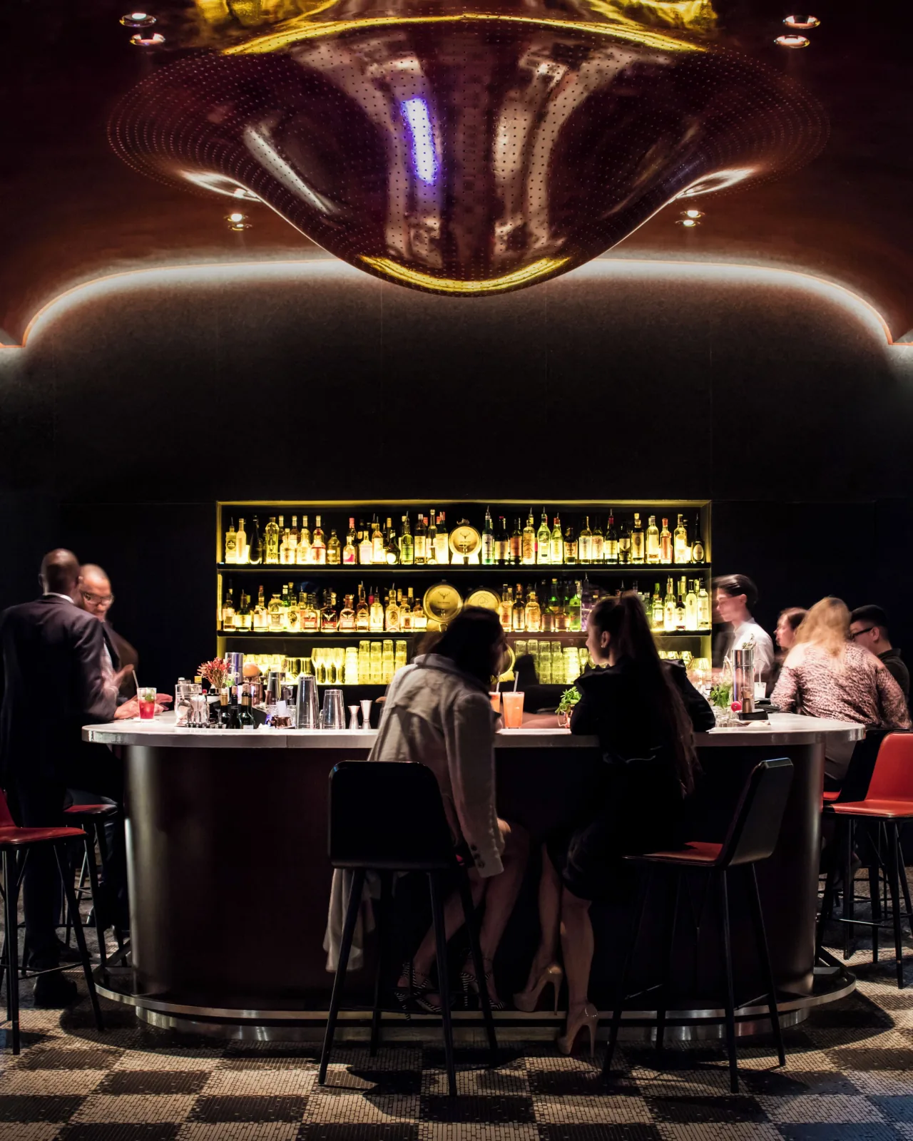 Guests at a round bar counter under a lacquered ceiling, shelves of lit bottles behind