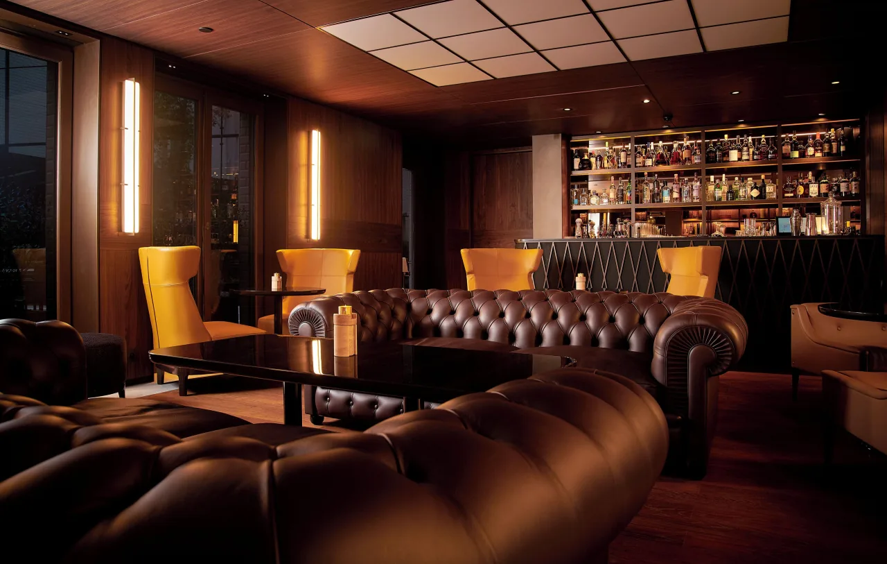 Un salon lambrissé sombre, canapés de cuir brun, fauteuils jaunes et un bar garni de bouteilles