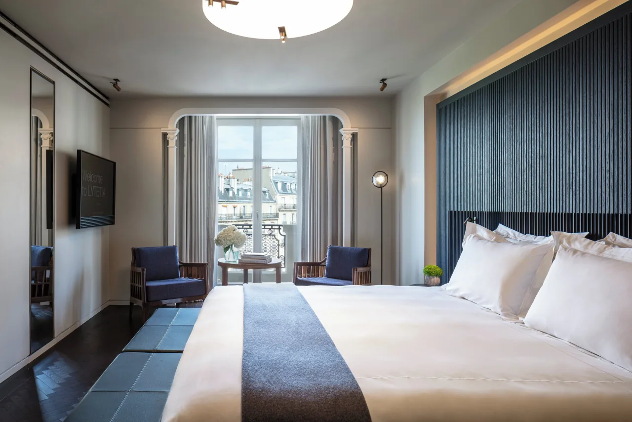Une chambre, grand lit blanc contre un mur de lattes bleu nuit, deux fauteuils bleus et une petite table face à une porte-fenêtre sur les immeubles de Paris