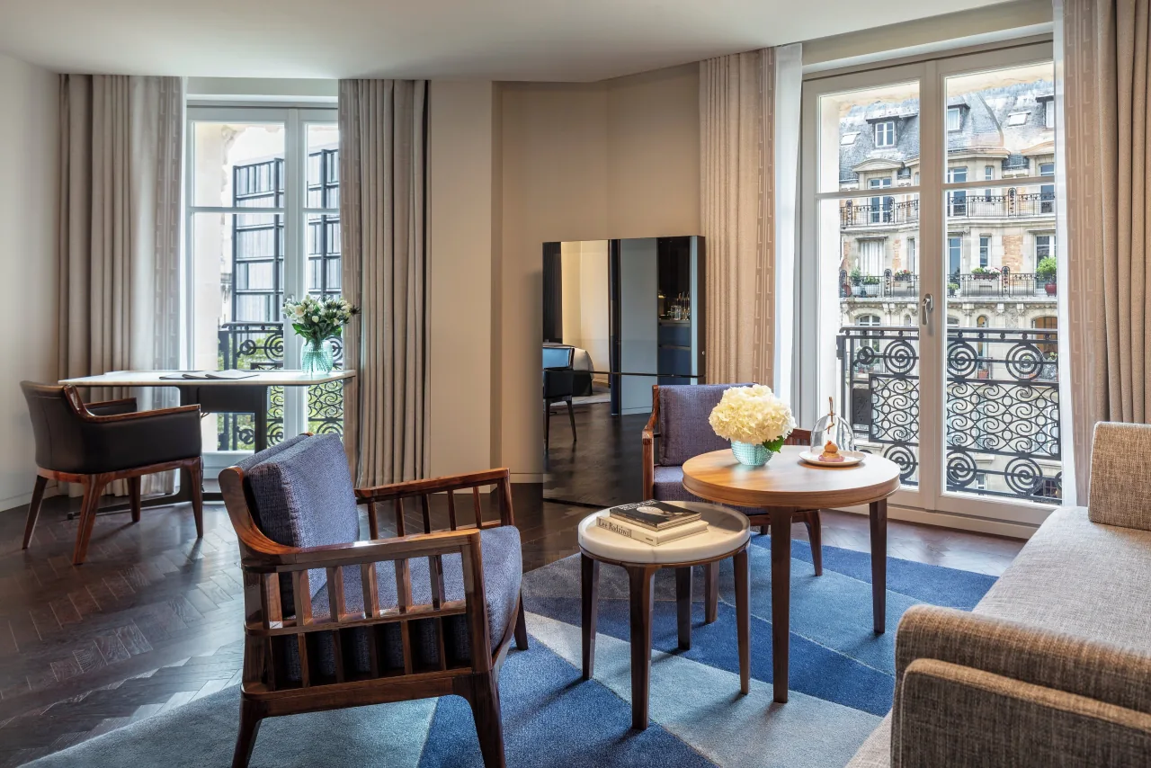 Un salon, fauteuils de bois, tables rondes et canapé sur un tapis bleu, portes-fenêtres à balcons de fer forgé face aux immeubles parisiens