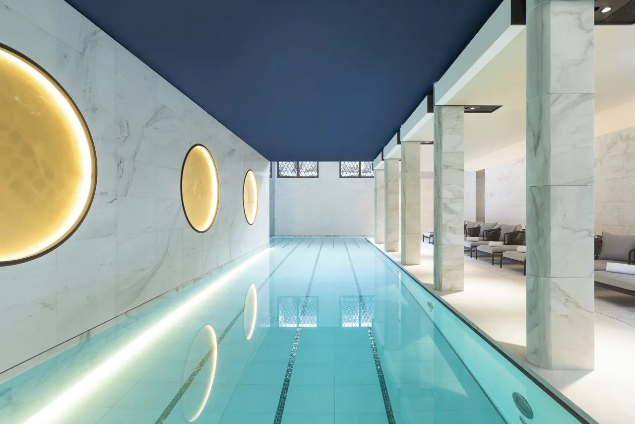 Une longue piscine intérieure entre des murs de marbre blanc ornés de disques dorés lumineux, sous un plafond bleu nuit, lignes d’eau et transats derrière une rangée de piliers de marbre
