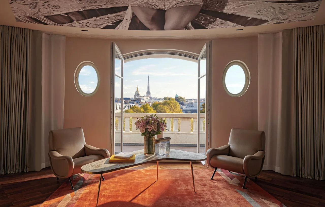 Un salon arrondi, deux oculus et une porte-fenêtre ouverte sur une balustrade de pierre, la tour Eiffel et le dôme des Invalides au loin, deux fauteuils de cuir sur un tapis orange