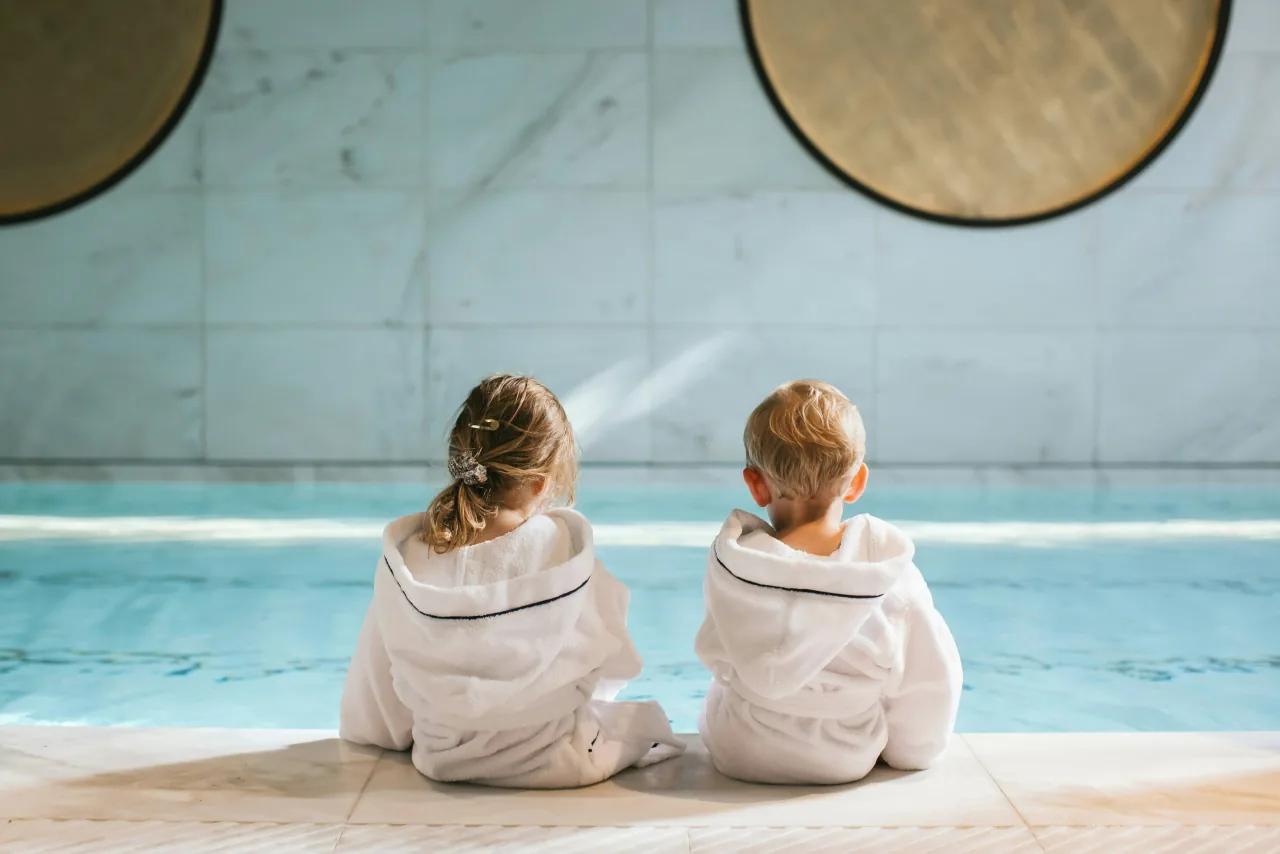 Deux enfants en peignoir blanc assis au bord de la piscine intérieure, vus de dos