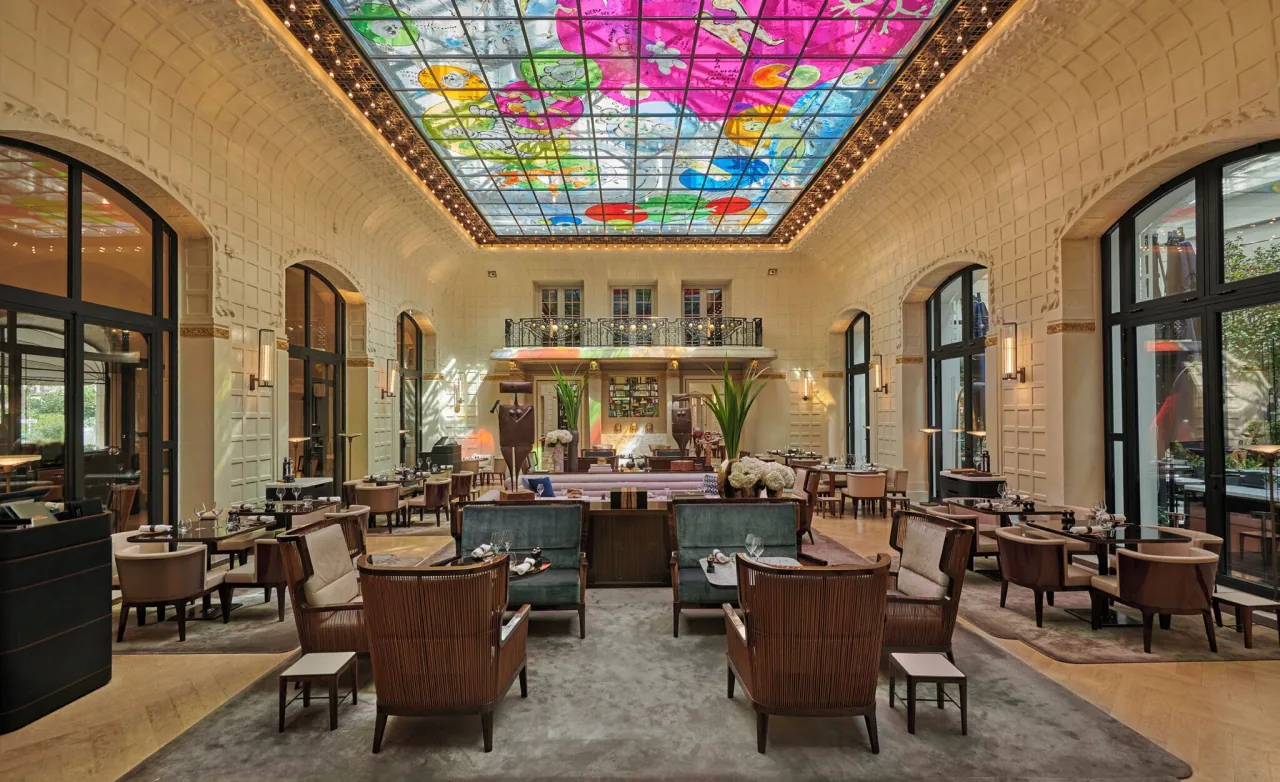 Un long salon sous une verrière peinte aux couleurs vives, fenêtres en arc et fauteuils autour de tables basses