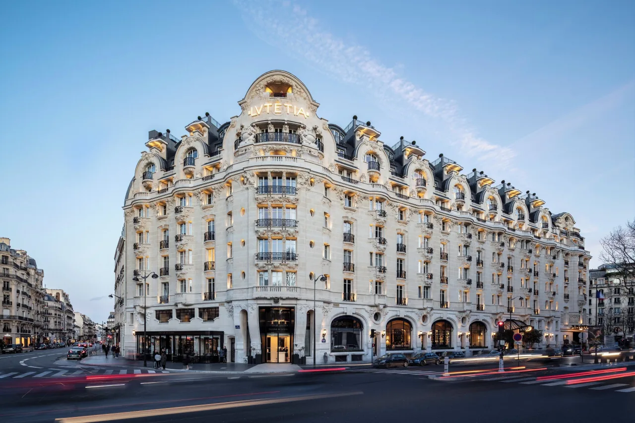 La façade Art nouveau de pierre claire de l’hôtel, à l’angle de deux rues au crépuscule, le nom LVTETIA au-dessus de l’angle arrondi, traînées de phares des voitures