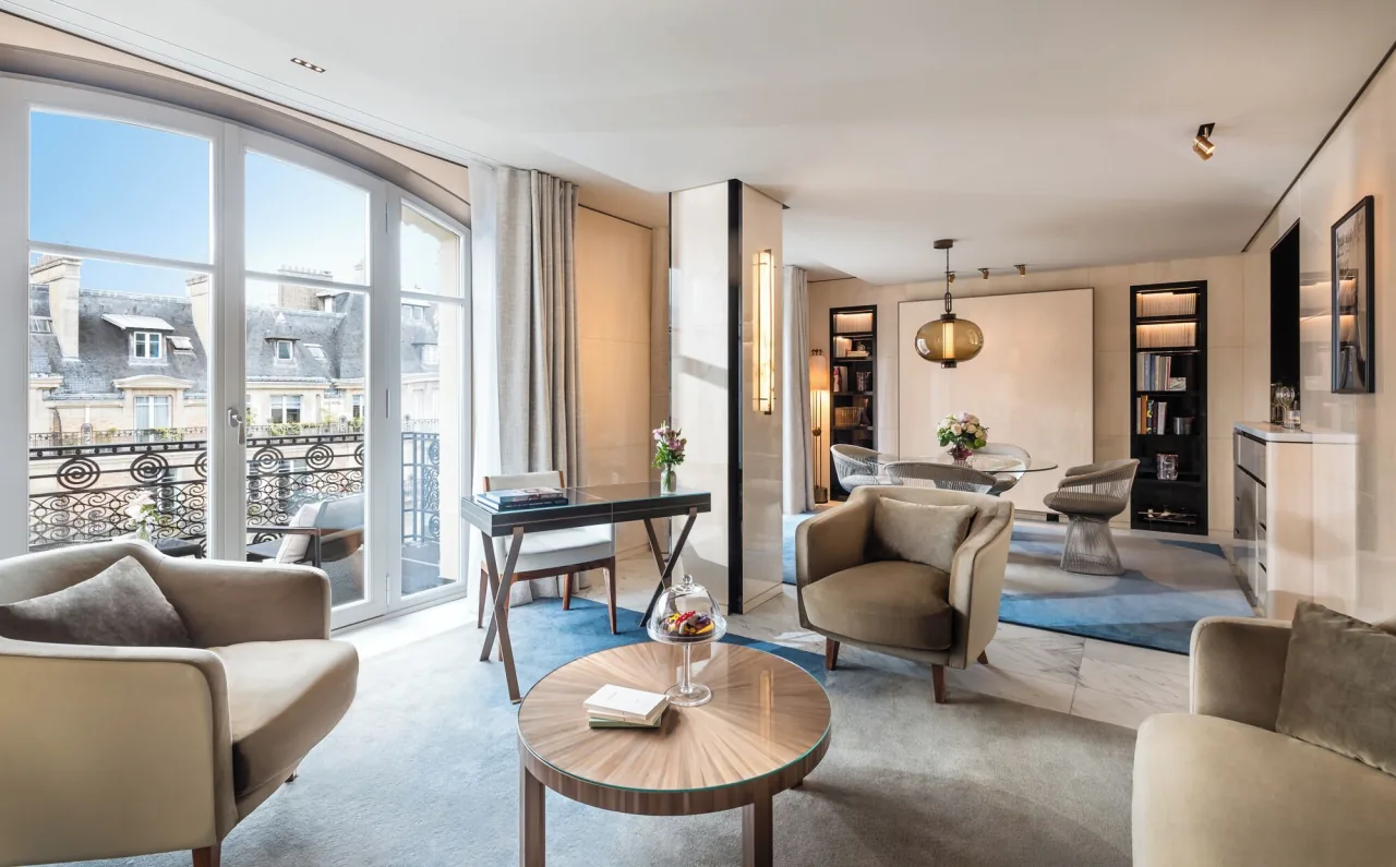 Un salon lumineux, hautes fenêtres sur un balcon de fer forgé, fauteuils clairs autour d’une table ronde en bois, un bureau et une table à manger