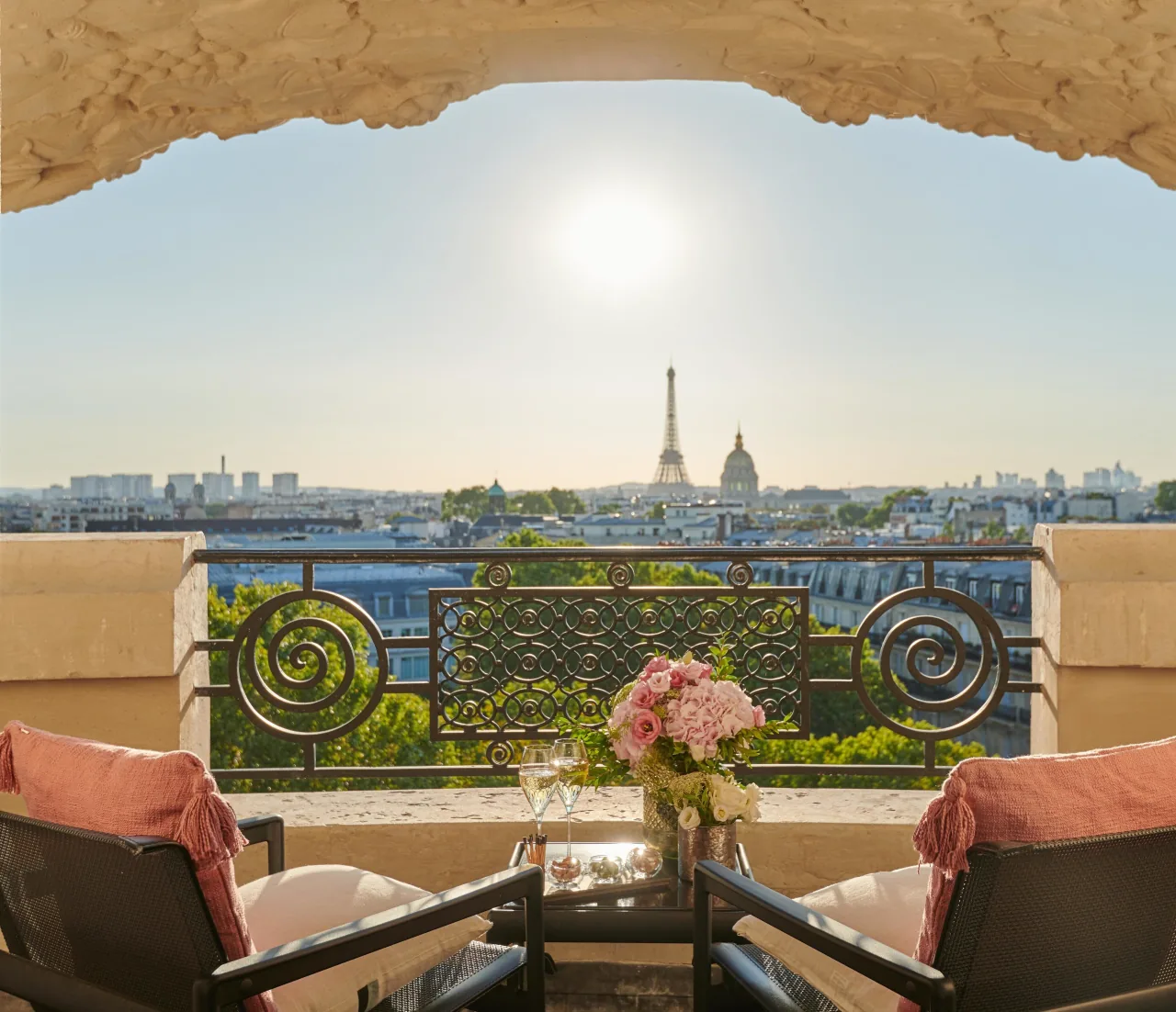 Un balcon sous une arche de pierre sculptée, deux fauteuils aux coussins roses, des roses et deux coupes, face aux toits de Paris, à la tour Eiffel et au dôme doré des Invalides