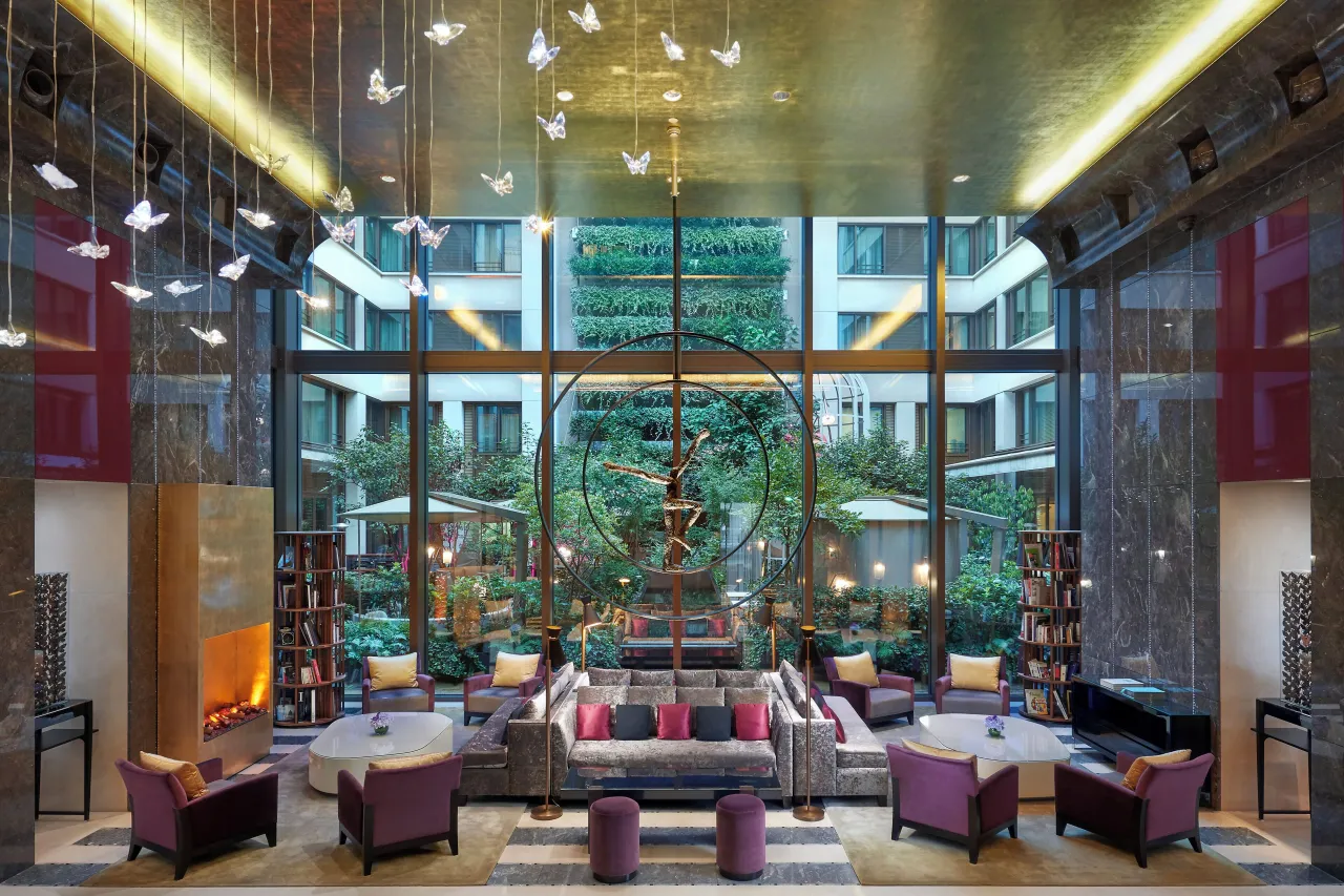 A lounge with plum armchairs and a fireplace, opening through tall windows onto a green courtyard garden, paper birds hanging from the ceiling