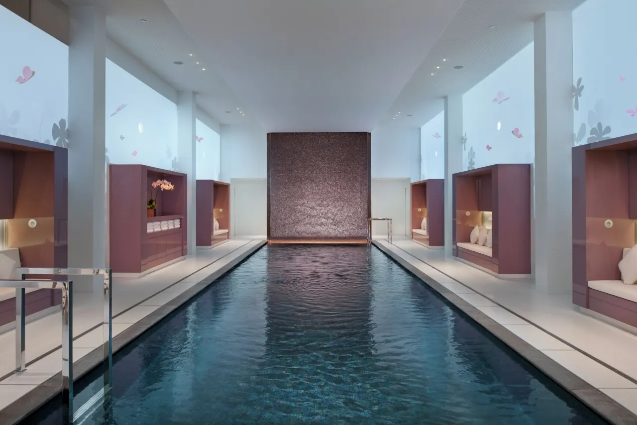 A long, dark indoor pool between pale columns, with plum-coloured alcoves holding cushions and a mosaic wall at the end