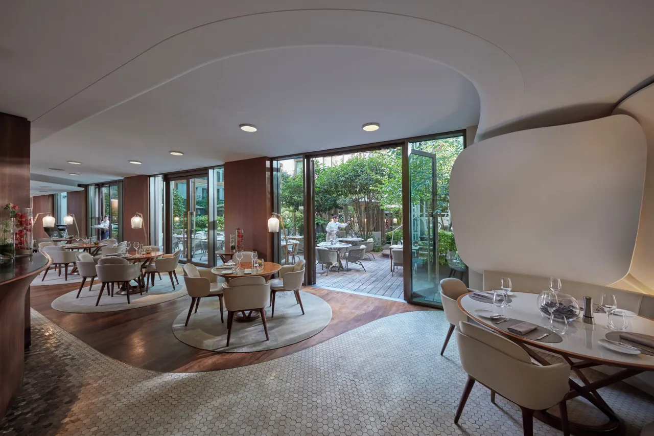 A restaurant with round tables, curved white walls and glass doors opening onto a planted terrace