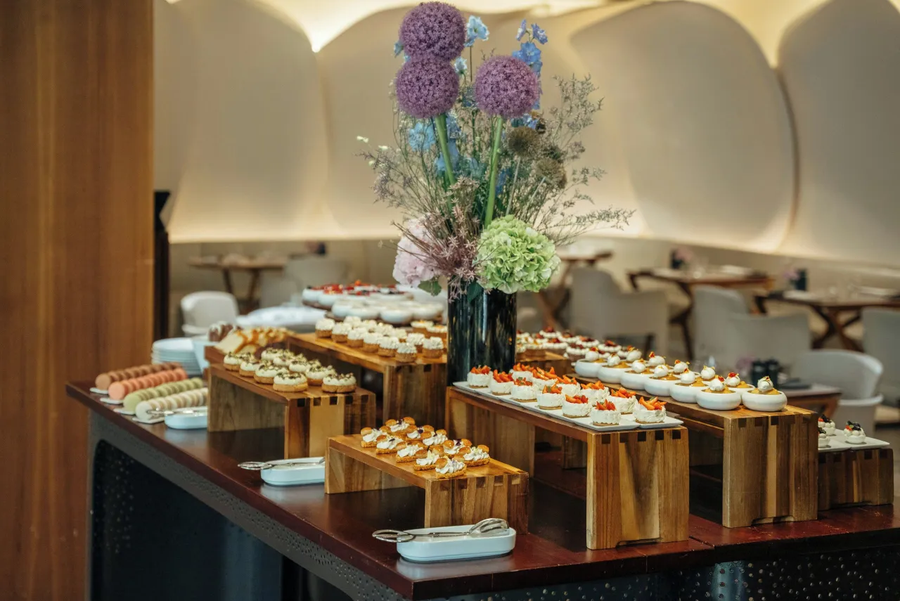 A pastry counter with rows of small cakes on wooden stands and a bouquet of alliums