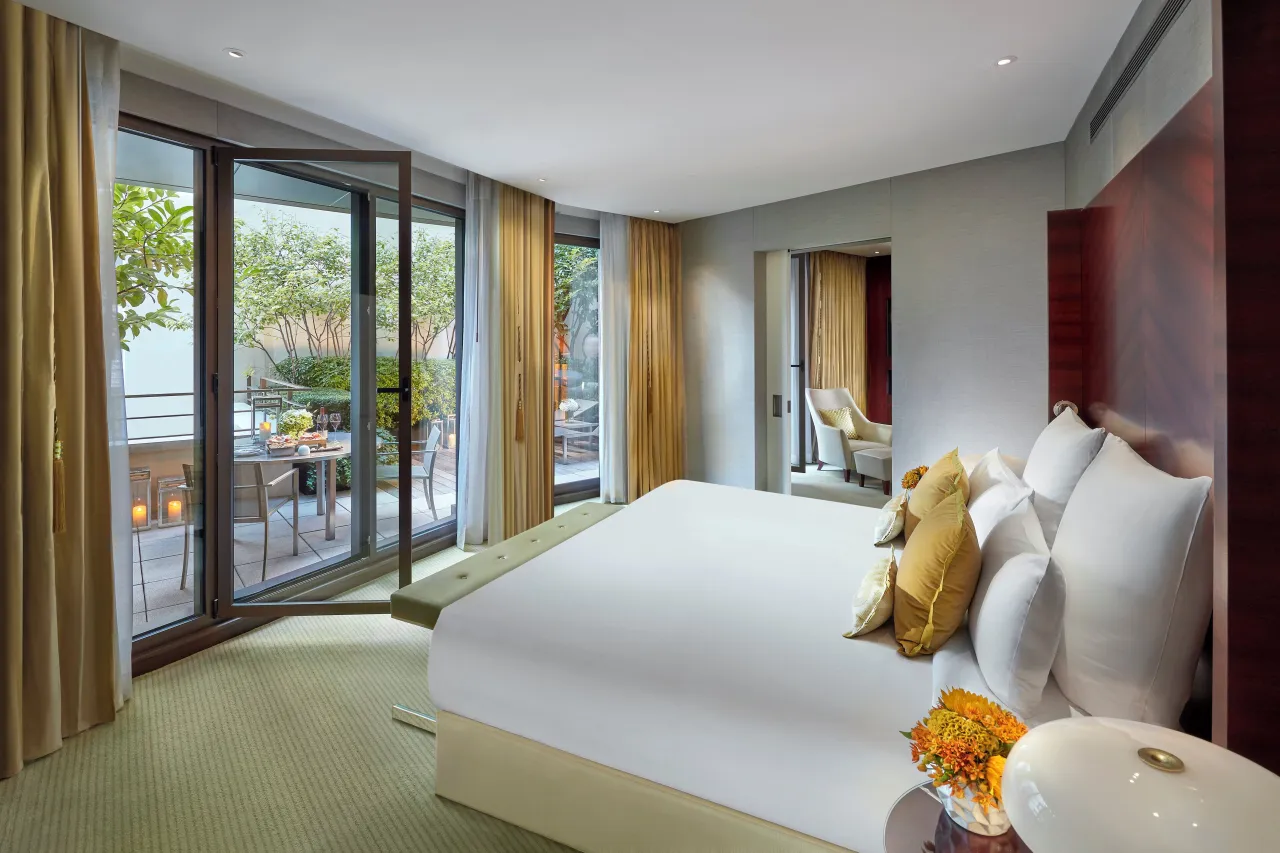 A bedroom with a large white bed and yellow cushions, French windows open onto a terrace with trees