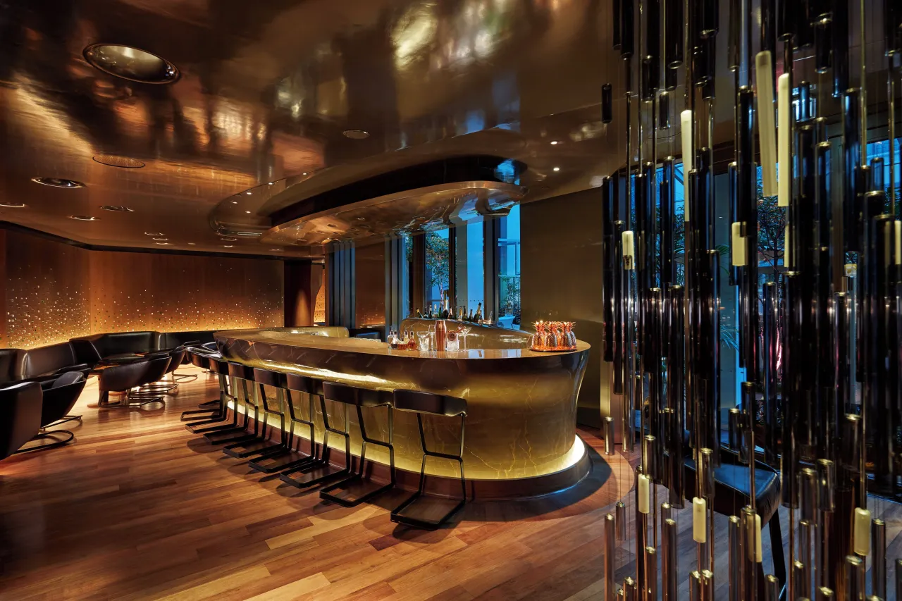 An oval bar under a gilded ceiling, with high stools, a wood floor and a screen of dark glass rods