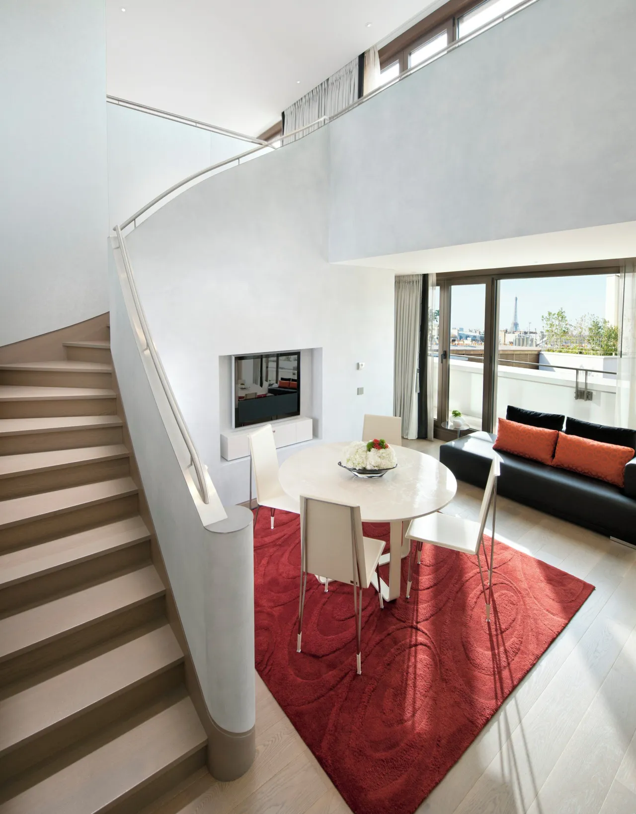 A curved white staircase in a two-level suite, a round dining table on a red rug and windows onto the rooftops