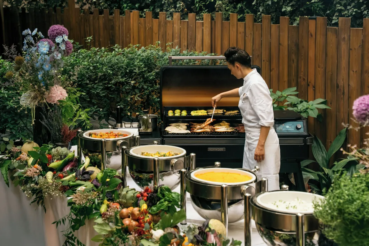 A cook at a barbecue in a garden with flowers, vegetables and copper chafing dishes