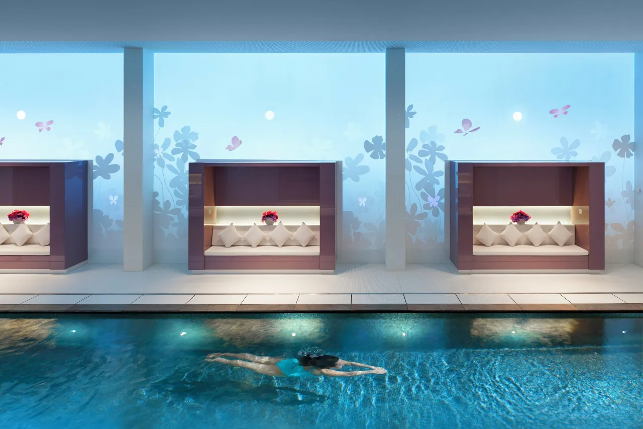 A swimmer in a lit blue pool, below windows printed with flowers and three cushioned alcoves