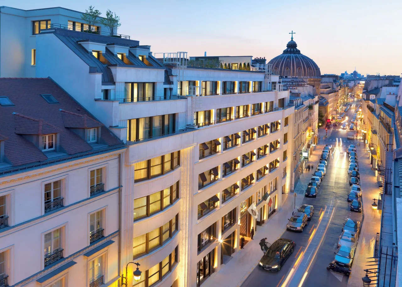 The hotel’s pale facade along a narrow street at dusk, with lit windows, parked cars and a dome at the end of the street