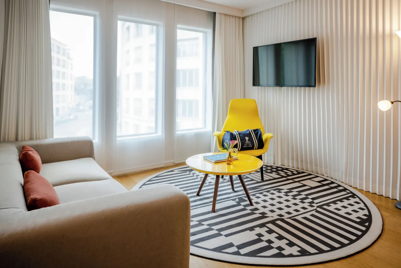 Un salon, un canapé gris, un fauteuil jaune, un tapis rond graphique et de hautes fenêtres
