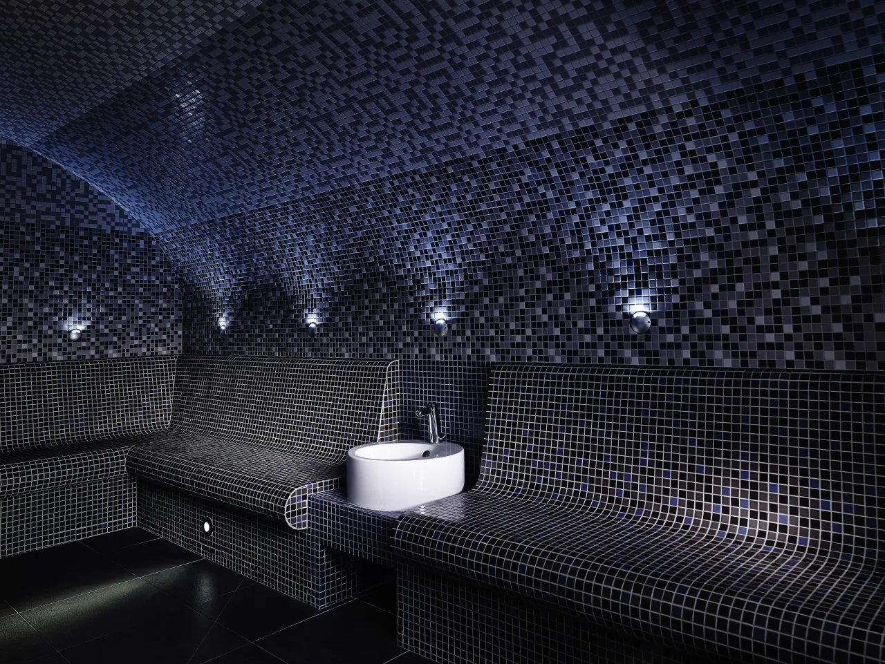 Un hammam en mosaïque sombre, une vasque de pierre et de petites lumières