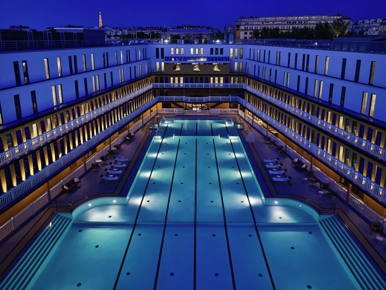 Le bassin extérieur la nuit, éclairé de bleu entre les étages blancs des balcons, la ville au fond