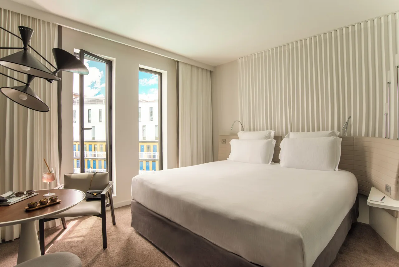 Une chambre, un lit blanc, des murs clairs nervurés, une applique articulée et de hautes fenêtres