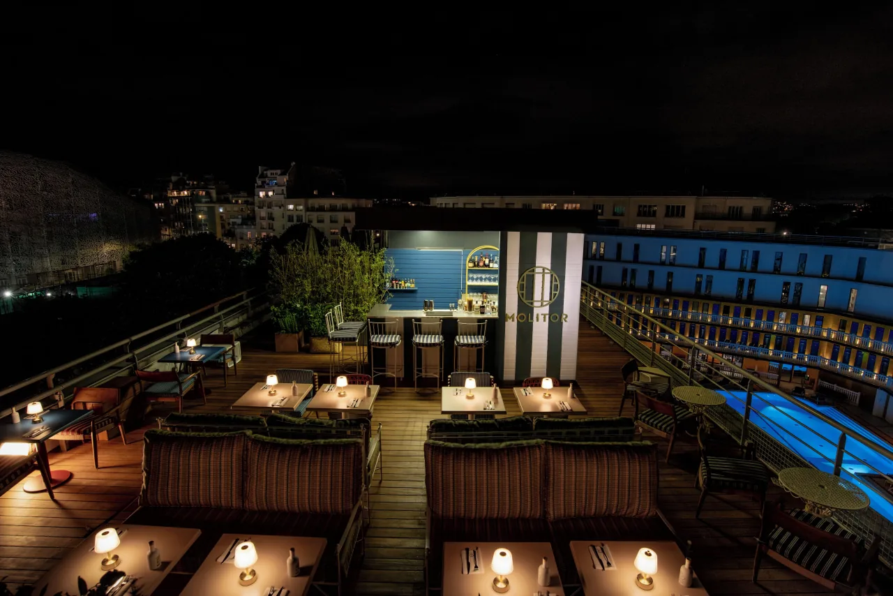 Un toit-terrasse la nuit, des tables basses et des lampes, le bassin éclairé en contrebas