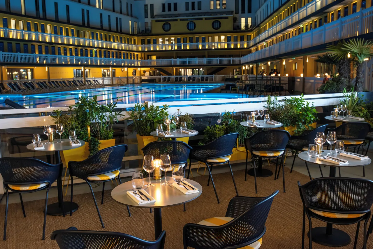 Des tables sur une terrasse au bord du bassin éclairé, le soir, les étages de cabines au fond