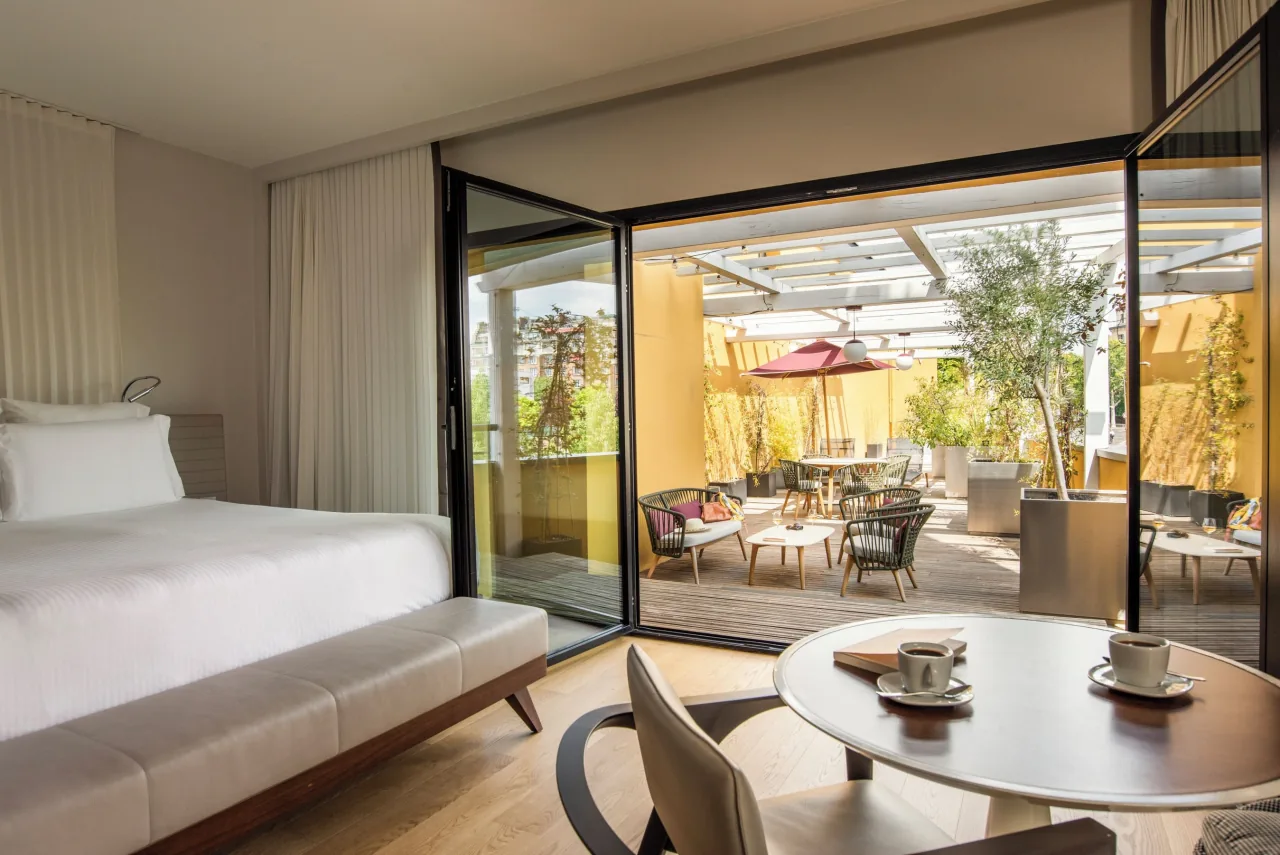 Une chambre ouverte par des portes vitrées sur une terrasse plantée, avec des tables et des parasols