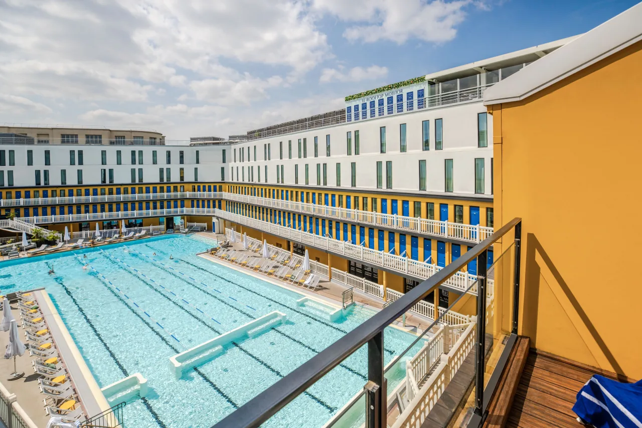 Un bassin extérieur bordé de portes de cabines bleues et de balustrades blanches sur trois niveaux, des transats et un bâtiment jaune à l’angle