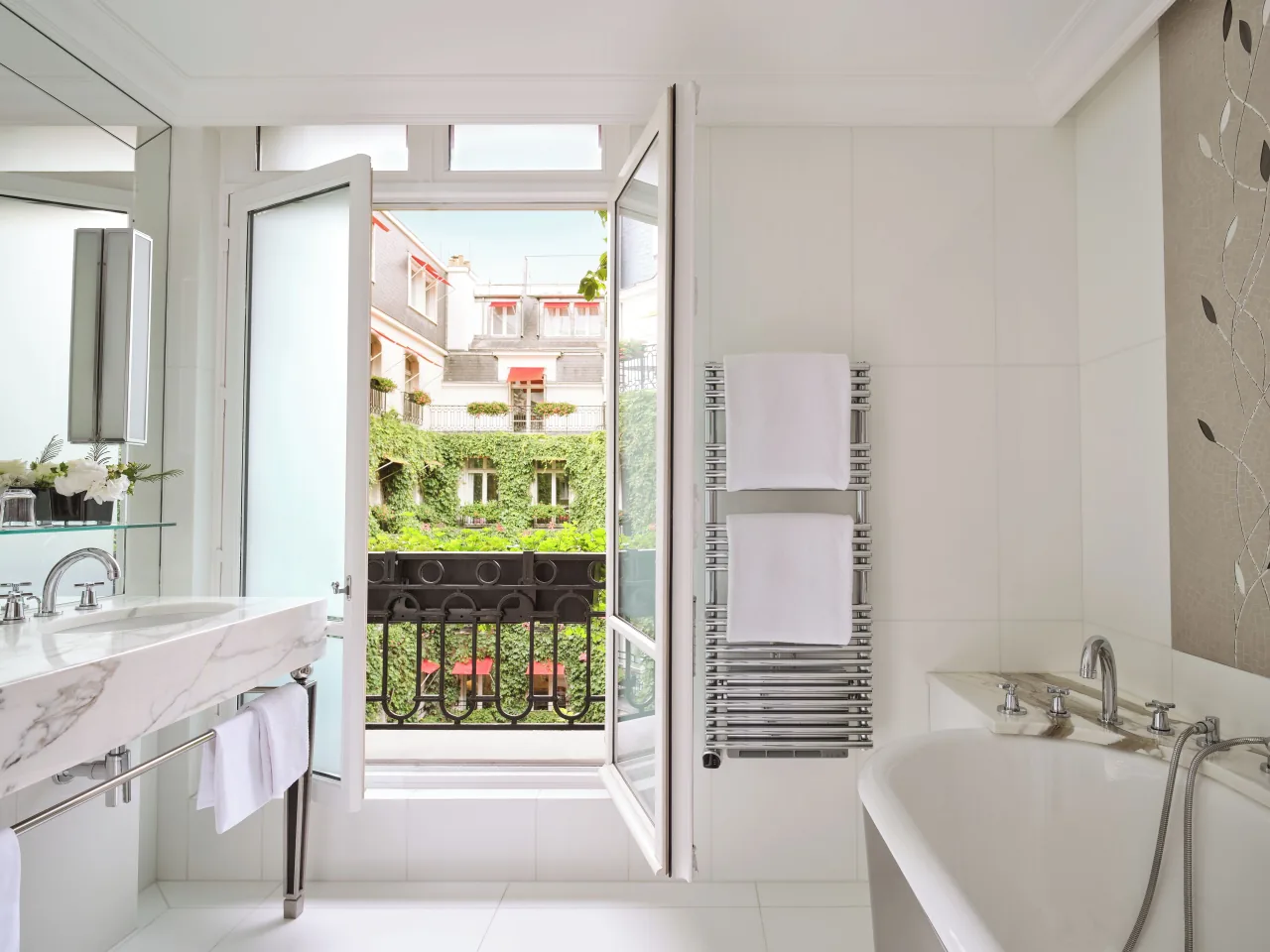A white marble bathroom with a washbasin, a bathtub and a towel rail, its French window open onto a balcony over the ivy-covered courtyard