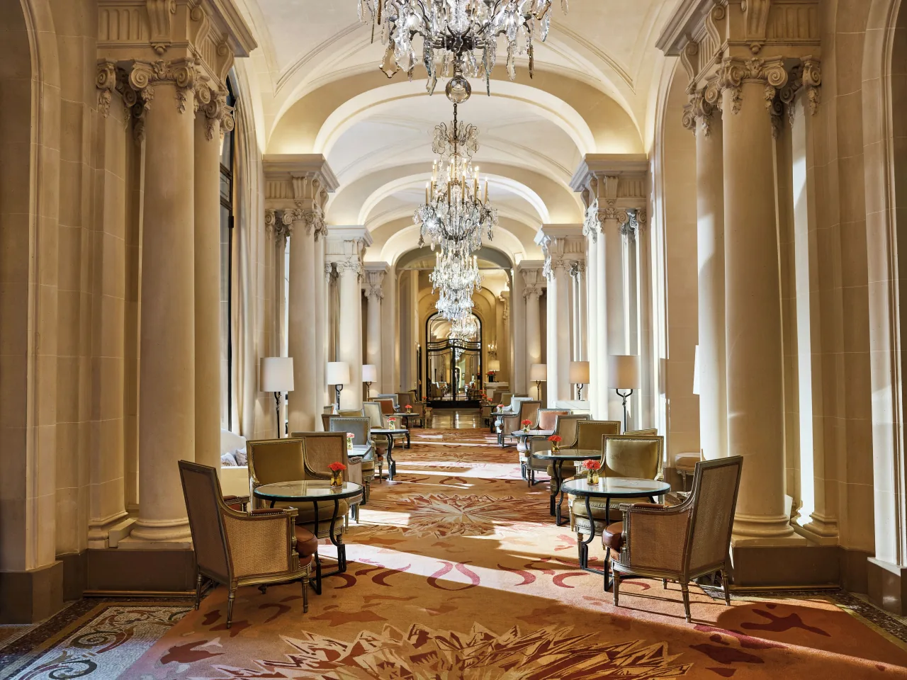 A long gallery of stone columns and arches lit by crystal chandeliers, with armchairs around small round tables on a patterned carpet
