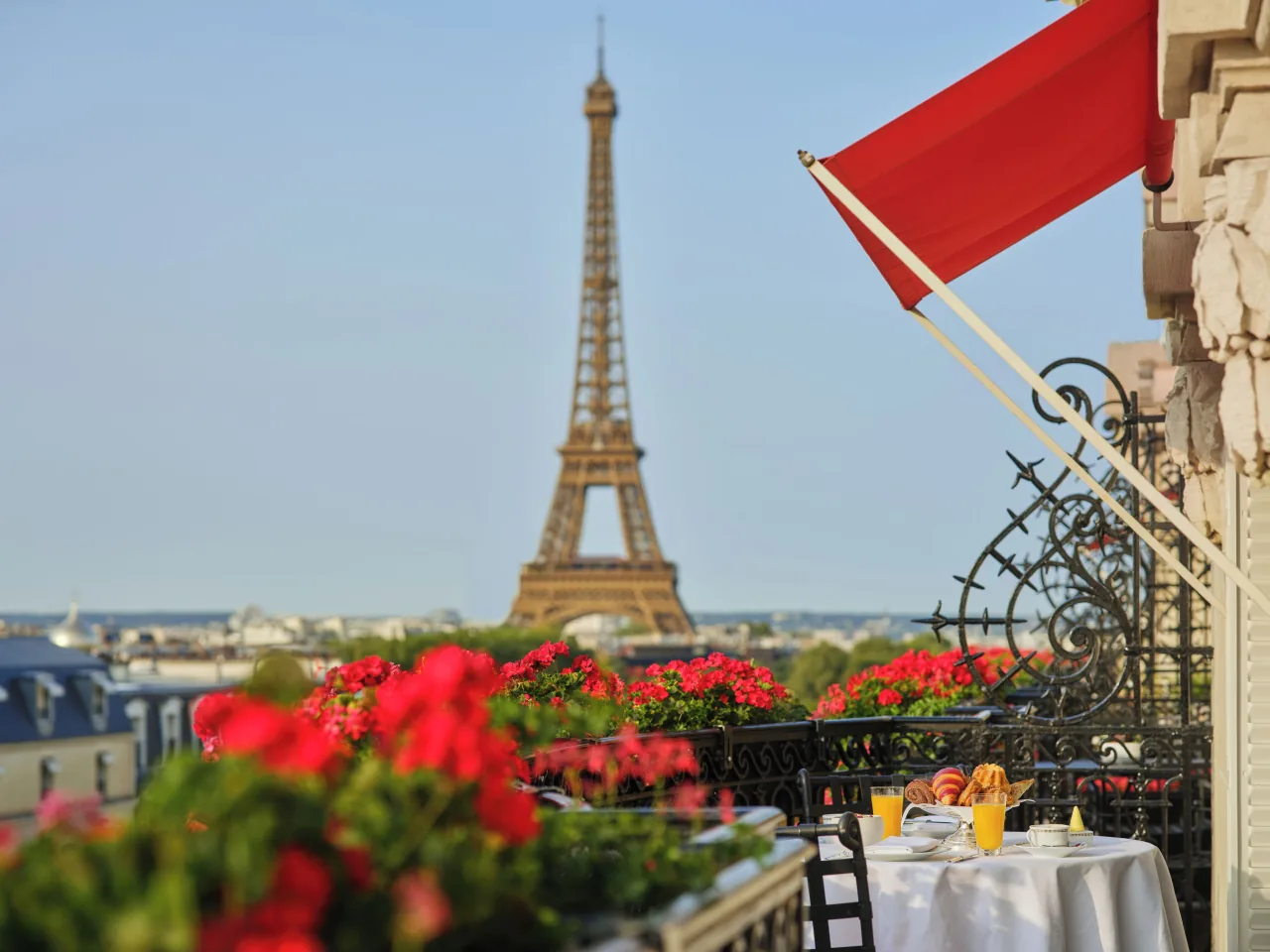 The Eiffel Tower seen from a balcony lined with red geraniums, under a red awning, beside a breakfast table set with orange juice